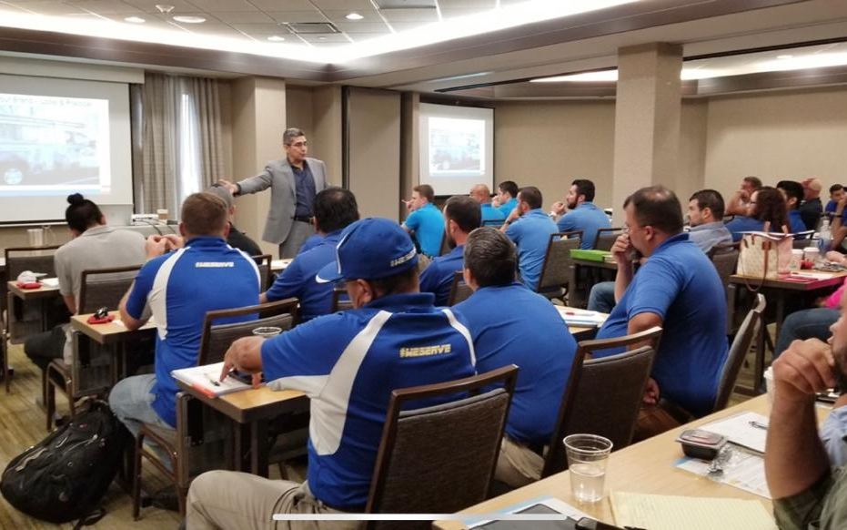 A hotel conference room full of roofing sales reps in matching blue polos during a training session
